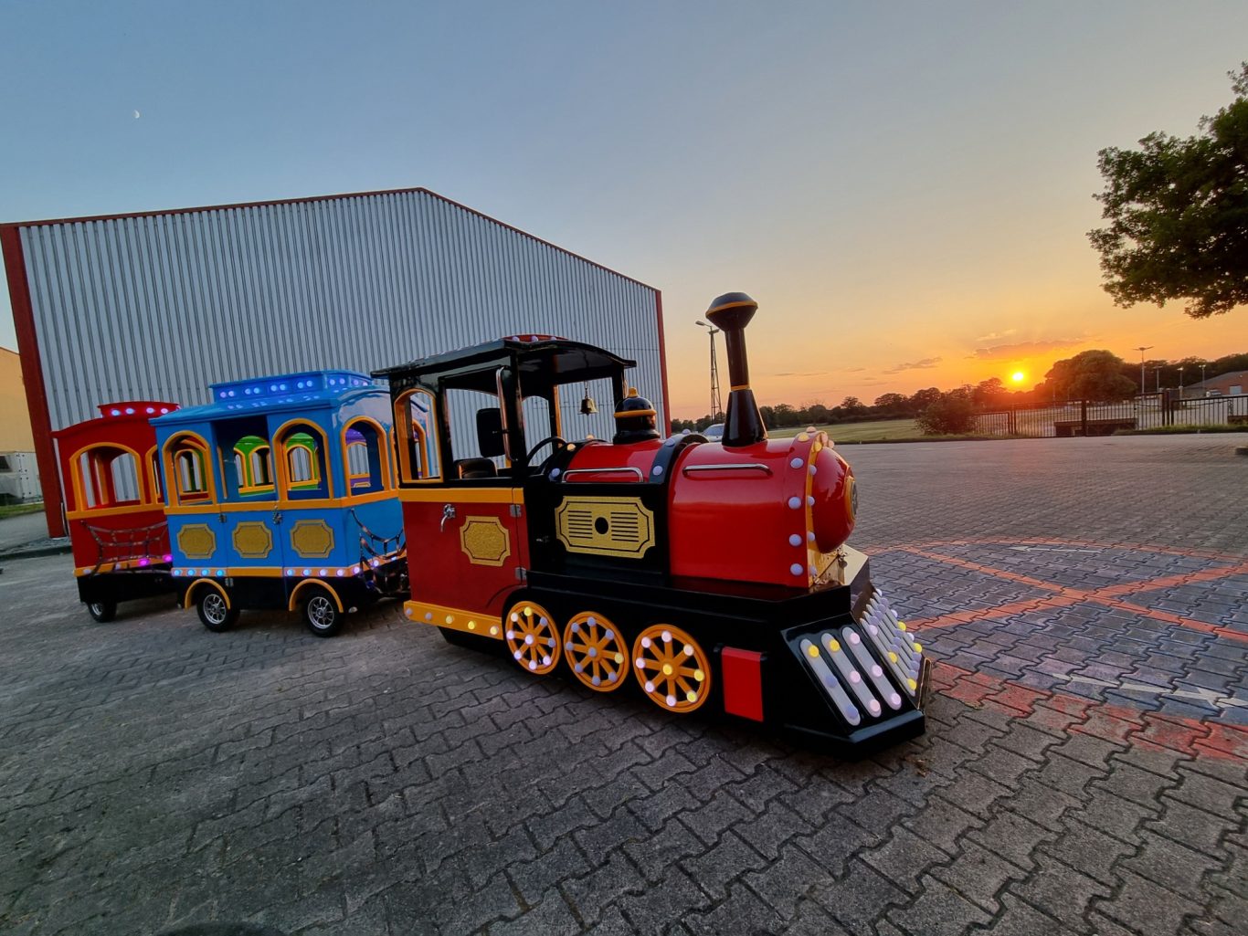 Bimmelbahn II Bunte Eisenbahnlok und Waggons stehen vor einem Sonnenuntergang auf einem Parkplatz.